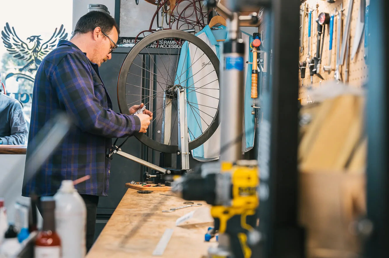 A mechanic in a plaid shirt works on a bicycle wheel held in a stand beside a tool-covered workbench.