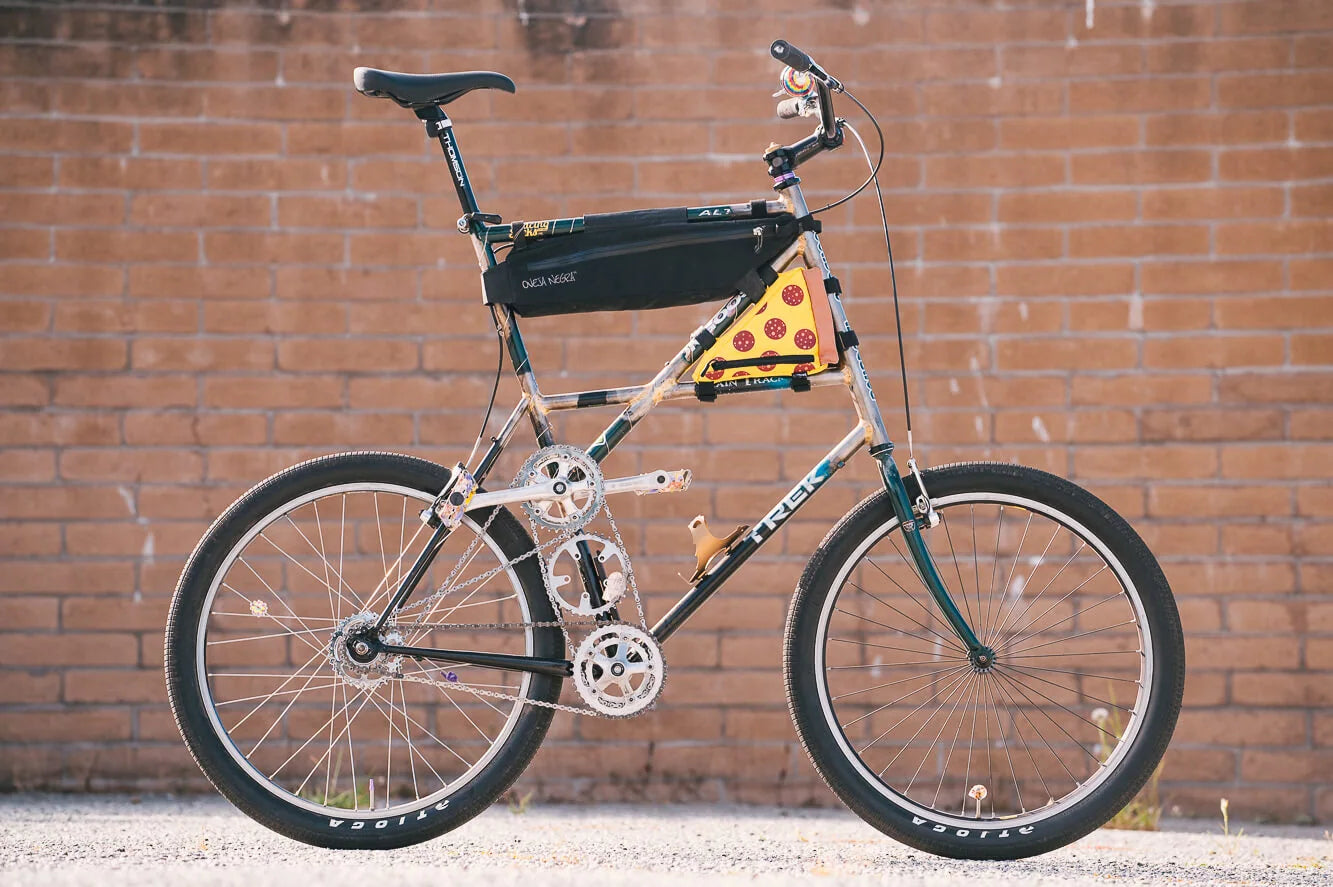 A tall bicycle with stacked frame sections, two chain levels, a black frame bag and a pizza-patterned pouch against a brick wall.
