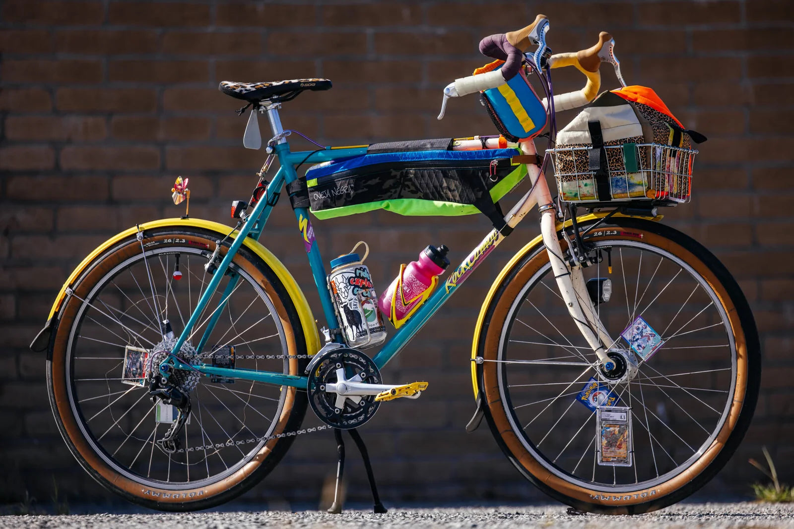 A turquoise-and-white drop-bar bicycle with yellow fenders, colorful bags, a loaded front basket and cards in the spokes.