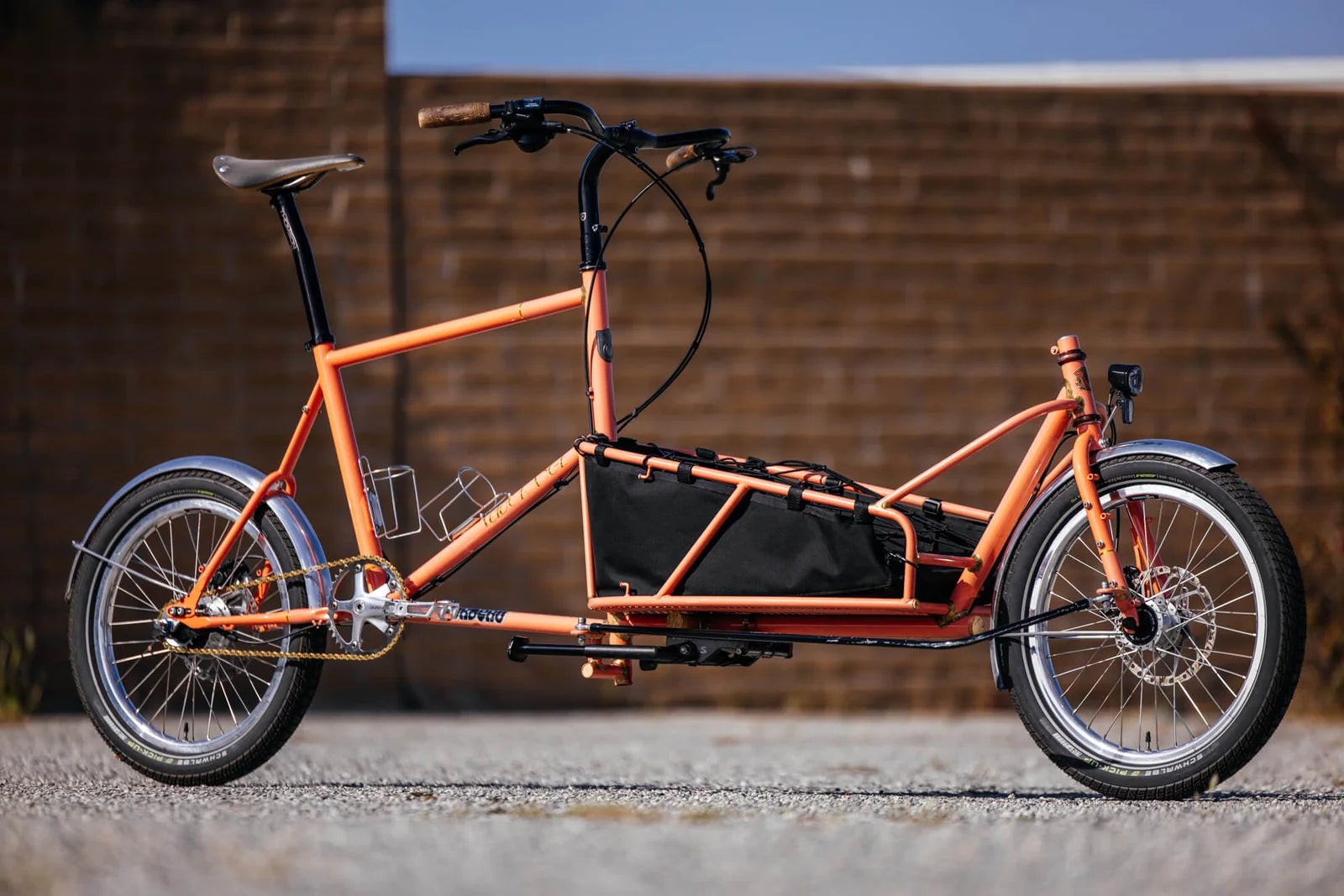 An orange cargo bicycle with a long low platform and black cargo bag, shown side-on against a brick wall.